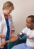A healthcare professional wearing a white coat and a stethoscope is measuring the blood pressure of a seated man. The man is wearing a casual t-shirt and light blue pants. The setting appears to be a clinical or healthcare environment.