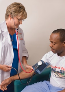 A healthcare professional wearing a white coat and a stethoscope is measuring the blood pressure of a seated man. The man is wearing a casual t-shirt and light blue pants. The setting appears to be a clinical or healthcare environment.