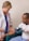 A healthcare professional wearing a white coat and a stethoscope is measuring the blood pressure of a seated man. The man is wearing a casual t-shirt and light blue pants. The setting appears to be a clinical or healthcare environment.