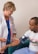 A healthcare professional wearing a white coat and a stethoscope is measuring the blood pressure of a seated man. The man is wearing a casual t-shirt and light blue pants. The setting appears to be a clinical or healthcare environment.