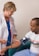 A healthcare professional wearing a white coat and a stethoscope is measuring the blood pressure of a seated man. The man is wearing a casual t-shirt and light blue pants. The setting appears to be a clinical or healthcare environment.