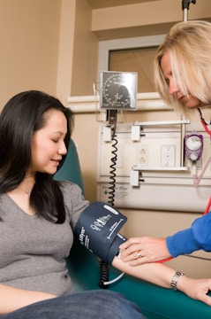 A healthcare professional measures a woman's blood pressure using a sphygmomanometer in a clinical setting. The woman sits on a green chair, and a medical device is seen attached to the wall in the background.