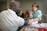 A healthcare professional measures the blood pressure of a man seated at a table. The man is wearing a white shirt while the healthcare worker is using a stethoscope and has a clipboard with papers. Brochures and red pens are visible on the table. Another person in a red shirt is seated in the background.