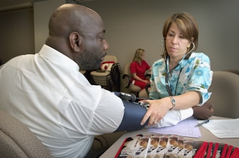 A healthcare professional measures the blood pressure of a man seated at a table. The man is wearing a white shirt while the healthcare worker is using a stethoscope and has a clipboard with papers. Brochures and red pens are visible on the table. Another person in a red shirt is seated in the background.