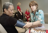 A healthcare professional is taking the blood pressure of a seated woman. The healthcare worker is wearing a stethoscope and a floral-patterned shirt, while the patient is wearing a dark shirt and sitting comfortably with her eyes closed. In the background, two women in red uniforms are engaged in conversation. Several pamphlets are visible on the table, suggesting a medical or educational setting.