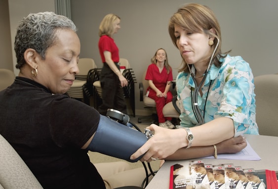 A healthcare professional is taking the blood pressure of a seated woman. The healthcare worker is wearing a stethoscope and a floral-patterned shirt, while the patient is wearing a dark shirt and sitting comfortably with her eyes closed. In the background, two women in red uniforms are engaged in conversation. Several pamphlets are visible on the table, suggesting a medical or educational setting.