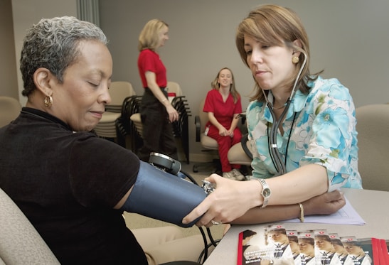 A healthcare professional is taking the blood pressure of a seated woman. The healthcare worker is wearing a stethoscope and a floral-patterned shirt, while the patient is wearing a dark shirt and sitting comfortably with her eyes closed. In the background, two women in red uniforms are engaged in conversation. Several pamphlets are visible on the table, suggesting a medical or educational setting.