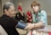 A healthcare professional is taking the blood pressure of a seated woman. The healthcare worker is wearing a stethoscope and a floral-patterned shirt, while the patient is wearing a dark shirt and sitting comfortably with her eyes closed. In the background, two women in red uniforms are engaged in conversation. Several pamphlets are visible on the table, suggesting a medical or educational setting.