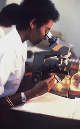 A person wearing a lab coat is using a microscope, seemingly focused and engaged in scientific observation. The setting suggests a laboratory environment with beakers and scientific tools visible in the background.