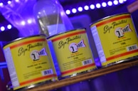 Three cans of '1 Shot' paint are prominently displayed on a shelf. The cans have distinctive yellow labels with bold text and a logo, set against a background featuring soft blue and purple lighting.