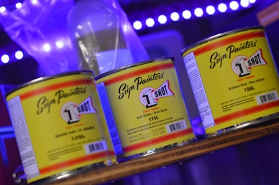 Three cans of '1 Shot' paint are prominently displayed on a shelf. The cans have distinctive yellow labels with bold text and a logo, set against a background featuring soft blue and purple lighting.