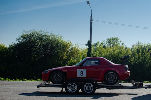A red sports car with a number 8 sticker on the door is situated on a trailer, parked on an open road. The car features a sporty design with visible modifications such as a rear spoiler and racing decals. It is set against a backdrop of lush green trees and a clear blue sky. A streetlight and power lines are visible in the distance.