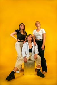 Three women are posing confidently against a bright yellow backdrop. The woman in the center is seated on a metallic box, wearing a striped shirt over a black top and beige pants, paired with black boots. The woman on the left stands with one hand on her hip, dressed in a black top and light-colored pants. The woman on the right wears a white top and black pants, with her hands resting at her sides. Each woman's expression exudes confidence and style.