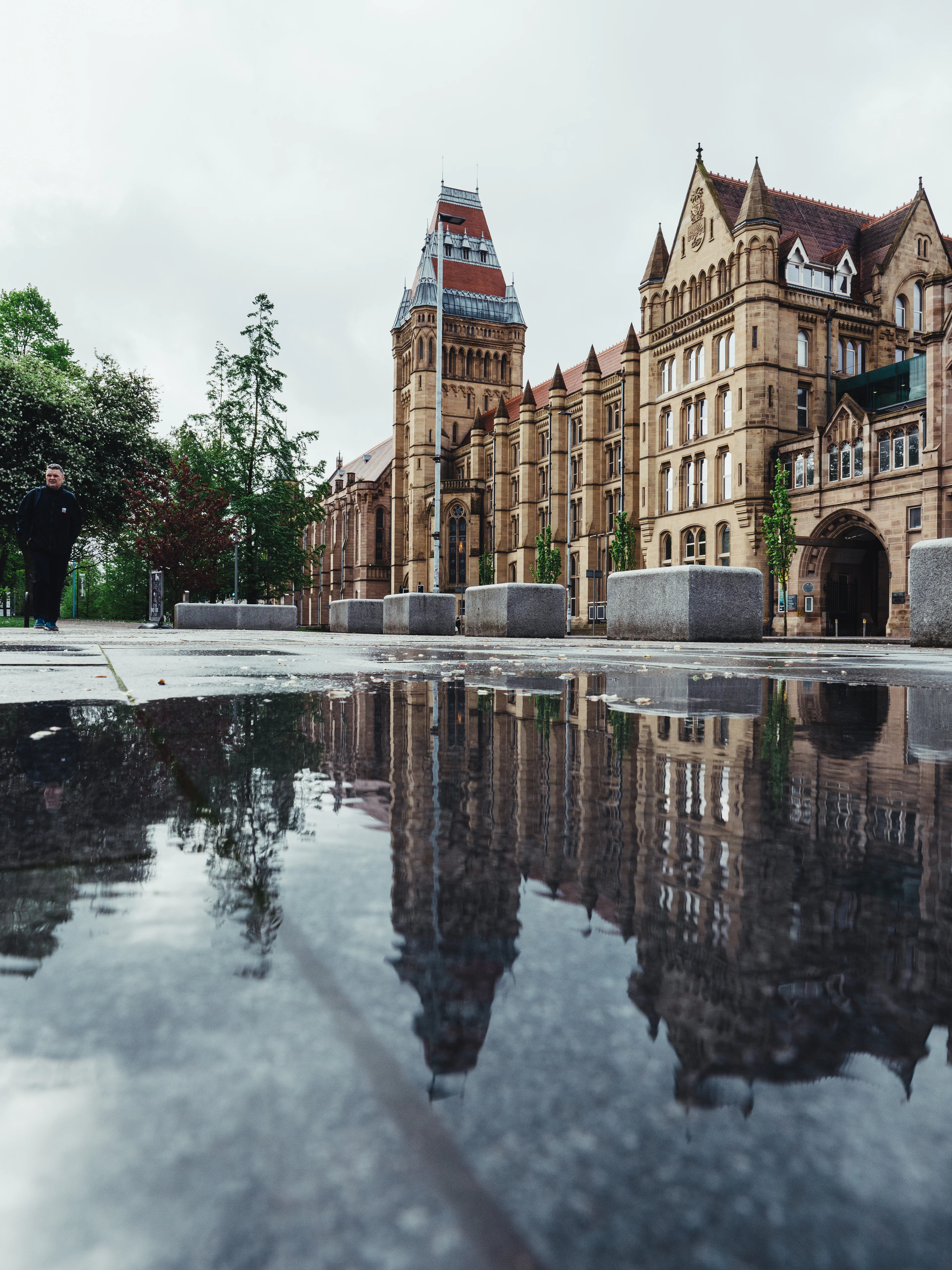 Gothic university buildings reflected in a wet plaza under an overcast sky