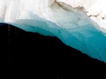 A massive iceberg with striking layers of blue and white ice, showing intricate textures and shades. The background contrasts sharply with the dark, shadowy void beneath the iceberg.