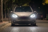 A car with bright headlights is parked on a dimly lit street at night. The illuminated surroundings create a focused atmosphere on the vehicle's front, where the license plate reads 'MAX VISION'. Trees and other parked cars are visible in the background.