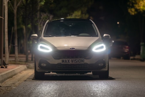 A car with bright headlights is parked on a dimly lit street at night. The illuminated surroundings create a focused atmosphere on the vehicle's front, where the license plate reads 'MAX VISION'. Trees and other parked cars are visible in the background.