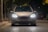 A car with bright headlights is parked on a dimly lit street at night. The illuminated surroundings create a focused atmosphere on the vehicle's front, where the license plate reads 'MAX VISION'. Trees and other parked cars are visible in the background.
