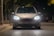 A car with bright headlights is parked on a dimly lit street at night. The illuminated surroundings create a focused atmosphere on the vehicle's front, where the license plate reads 'MAX VISION'. Trees and other parked cars are visible in the background.