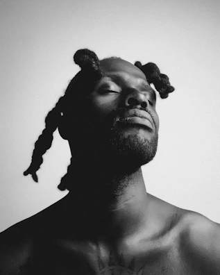 A black and white portrait of a person with closed eyes, showcasing textured hair styled in multiple sections. The lighting creates strong shadows, emphasizing facial features and the serene expression.
