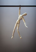 A wooden mannequin figure hangs from a horizontal rod with one hand, creating a sense of tension and imbalance. The figure's limbs are jointed, typical of an artist's mannequin, and are positioned dynamically as if in motion.