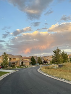 A suburban neighborhood with several houses lining a curved street, bordered by manicured lawns and trees. Above, the sky is filled with clouds, with some highlighted in a warm, orange-pink hue as if during sunset or sunrise.