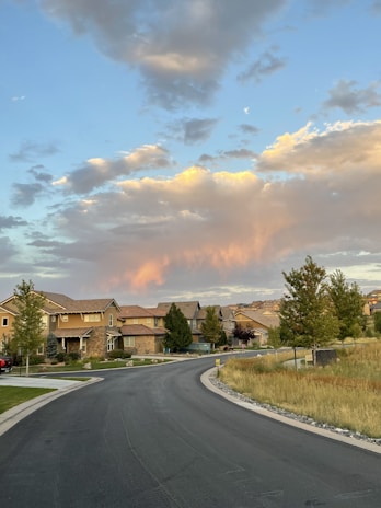 A suburban neighborhood with several houses lining a curved street, bordered by manicured lawns and trees. Above, the sky is filled with clouds, with some highlighted in a warm, orange-pink hue as if during sunset or sunrise.
