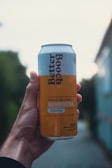 A hand holds a can of Better Booch kombucha with a label indicating it is a Golden Pear flavor. The can has a light blue and yellow design with text detailing the ingredients. The background is blurred, suggesting an outdoor setting with natural and urban elements.