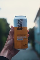 A hand holds a can of Better Booch kombucha with a label indicating it is a Golden Pear flavor. The can has a light blue and yellow design with text detailing the ingredients. The background is blurred, suggesting an outdoor setting with natural and urban elements.