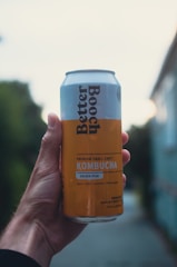 A hand holds a can of Better Booch kombucha with a label indicating it is a Golden Pear flavor. The can has a light blue and yellow design with text detailing the ingredients. The background is blurred, suggesting an outdoor setting with natural and urban elements.