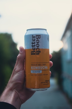 A hand holds a can of Better Booch kombucha with a label indicating it is a Golden Pear flavor. The can has a light blue and yellow design with text detailing the ingredients. The background is blurred, suggesting an outdoor setting with natural and urban elements.