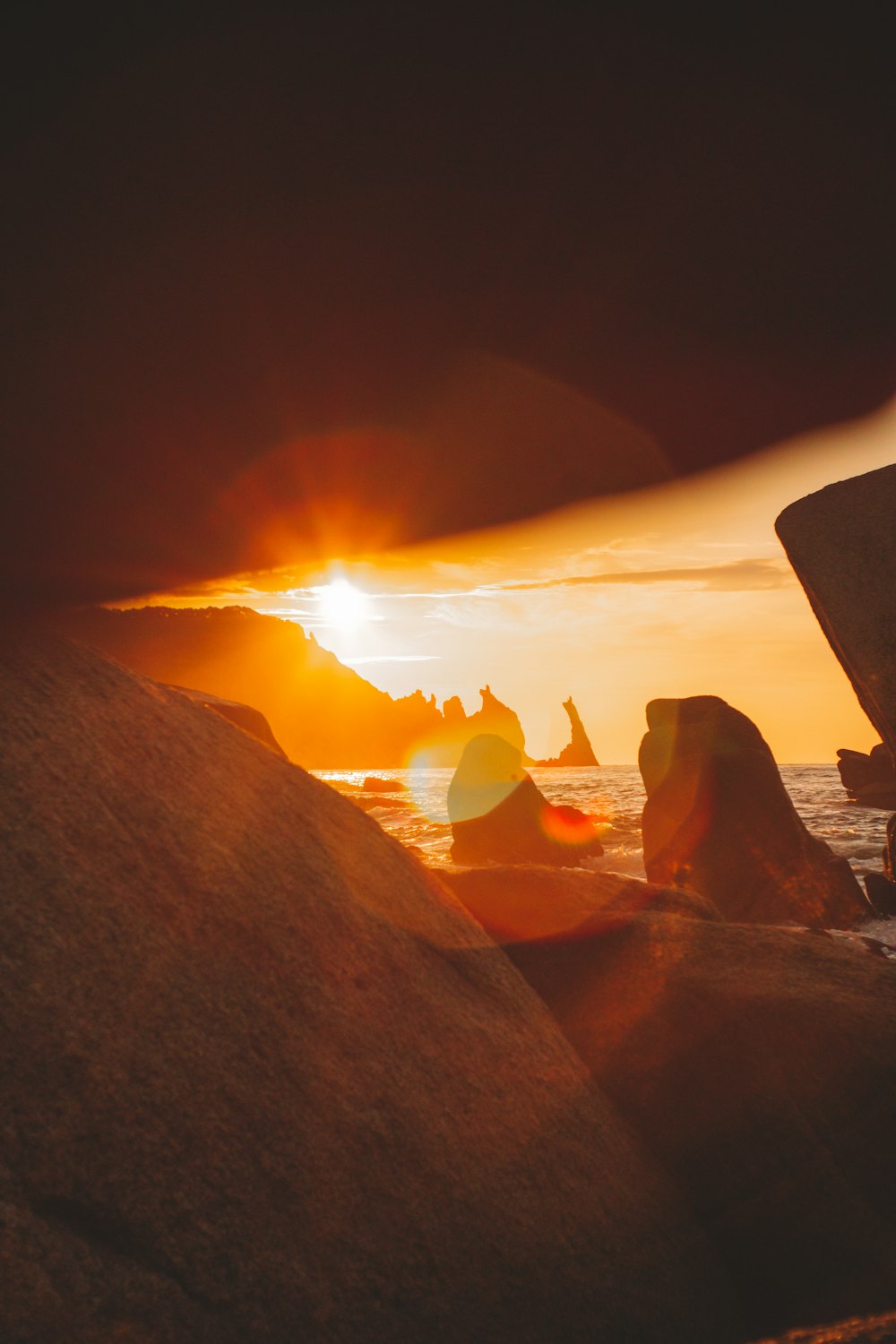 the sun is setting over the ocean with rocks in the foreground