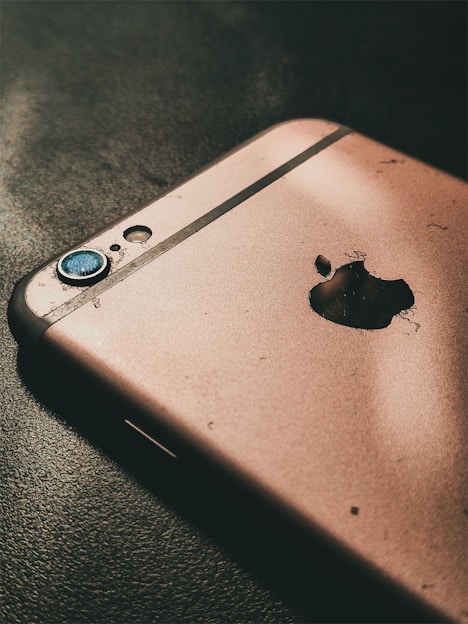 A close-up of a damaged smartphone with a focus on its rear side, showing a cracked and worn-out surface. The camera lens is scratched and there is an evident broken area near the manufacturer's logo.
