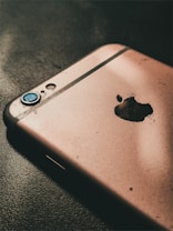 A close-up of a damaged smartphone with a focus on its rear side, showing a cracked and worn-out surface. The camera lens is scratched and there is an evident broken area near the manufacturer's logo.