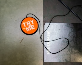 A round orange button with the text 'TRY ME' in white letters sits on a flat surface. A black cord is attached to the button and loops around on the surface. To the right of the button, part of a poster or sign with the text 'Grave Warrior' is visible. The surface appears textured and gray.