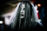 A ghostly figure with a cracked, white mask covered by a dark, translucent veil. The background is blurred, creating an eerie and mysterious atmosphere.