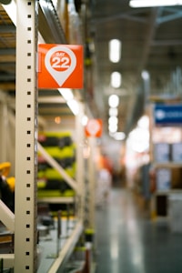 A long aisle in a warehouse or large retail store features shelves lined with various products. An orange sign with the text 'Bay 22' is prominently displayed on the left side. The background is slightly blurred, with soft lighting and distant shelving visible.