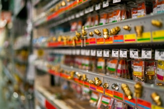 Rows of metal fittings and adapters are neatly arranged on display shelves in a store. Each item is attached to color-coded labels and price tags, giving a sense of organization and variety. The shelves extend into the background, suggesting a large selection of hardware items.