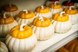 Several ceramic pumpkin-shaped containers with white bases and yellow lids are neatly arranged on a shelf. The containers have a glossy finish and resemble small pumpkins with stems on top.