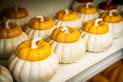 Several ceramic pumpkin-shaped containers with white bases and yellow lids are neatly arranged on a shelf. The containers have a glossy finish and resemble small pumpkins with stems on top.