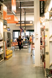 A bustling retail store environment with several people pushing shopping carts. Prominent signage for checkout and plumbing is visible above, and a visible display of various household products is on the shelves. The store has wide aisles and a bright interior.