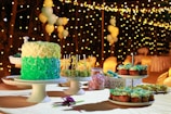 A richly decorated cake with layers of green and cream frosting is displayed on a cake stand surrounded by an assortment of desserts including cupcakes and cake pops. Soft lighting and a background adorned with strings of fairy lights and balloons create an elegant and festive atmosphere.