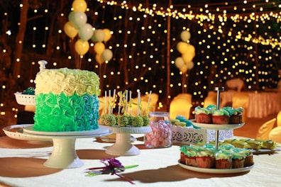 A richly decorated cake with layers of green and cream frosting is displayed on a cake stand surrounded by an assortment of desserts including cupcakes and cake pops. Soft lighting and a background adorned with strings of fairy lights and balloons create an elegant and festive atmosphere.