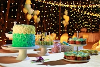 A richly decorated cake with layers of green and cream frosting is displayed on a cake stand surrounded by an assortment of desserts including cupcakes and cake pops. Soft lighting and a background adorned with strings of fairy lights and balloons create an elegant and festive atmosphere.