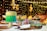A richly decorated cake with layers of green and cream frosting is displayed on a cake stand surrounded by an assortment of desserts including cupcakes and cake pops. Soft lighting and a background adorned with strings of fairy lights and balloons create an elegant and festive atmosphere.