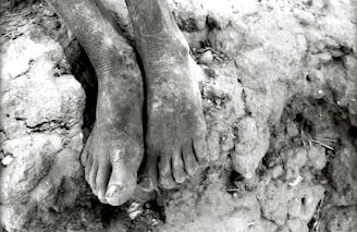 A pair of bare, dusty feet resting on rough, cracked earth. The texture of the skin and ground is emphasized, with visible cracks and dirt.