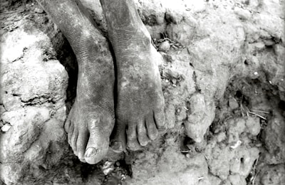 A pair of bare, dusty feet resting on rough, cracked earth. The texture of the skin and ground is emphasized, with visible cracks and dirt.