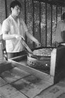 A person wearing a light shirt is cooking on a large stove. The setting appears to be indoors with perforated walls that let in natural light. The stove is large and round, suggesting it is meant for making a substantial dish.