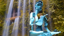 A contemporary metal sculpture of a seated figure appears amidst cascading waterfalls. The sculpture, painted in blue, has an abstract design with fragmented and angular features. Lush green foliage in the background adds a natural setting to the scene.