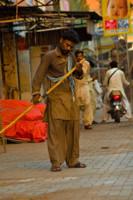 A man wearing traditional attire is sweeping the street with a broom. He is standing on a paved path surrounded by market stalls and blurred passersby in the background. There is a red covered object on the side and a person on a motorcycle in motion.